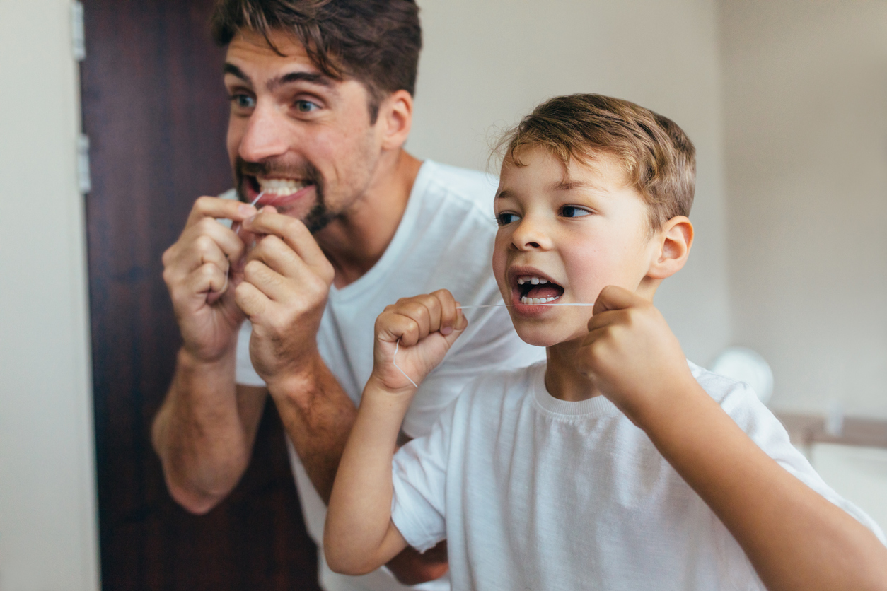 Little boy with his father in bathroom cleaning teeth with dental floss. Both looking in mirror and brushing teeth.